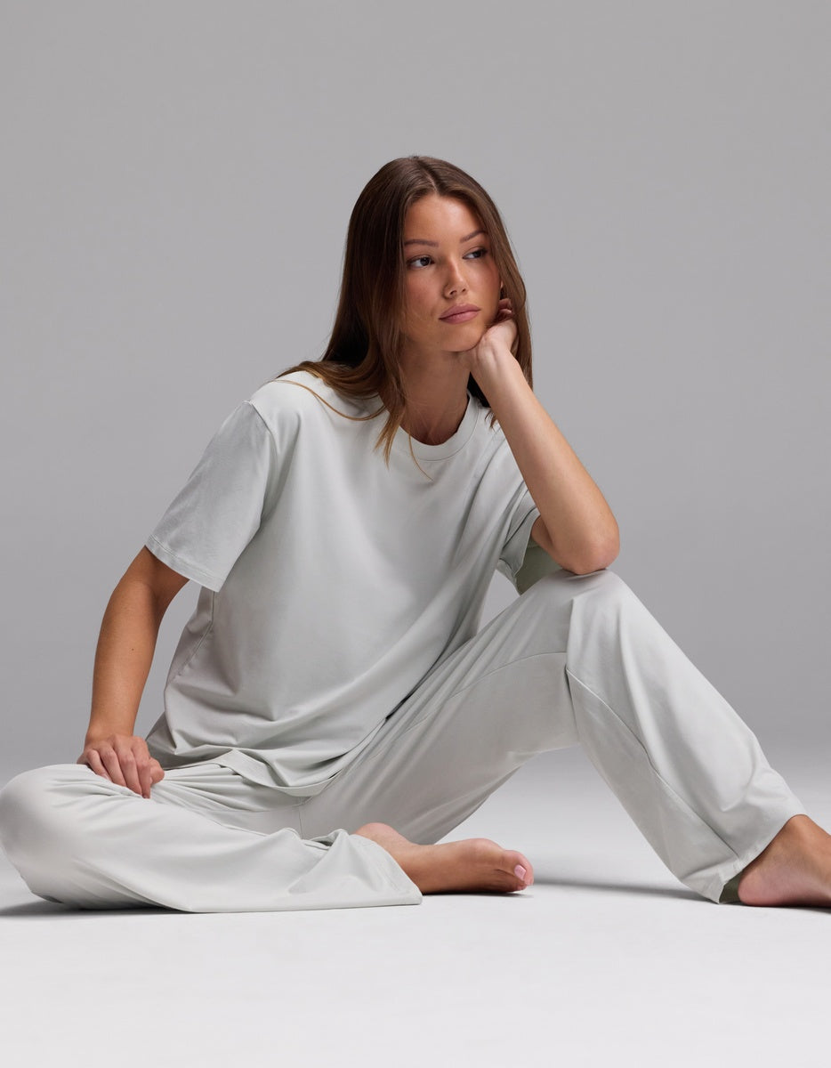Wearing the Cozy Earth Women’s First Layer Pajama Short Sleeve Tee & Pajama Pant Set in light gray, a woman sits barefoot on the floor against a plain gray background, one arm resting on her knee and a thoughtful expression on her face.|Color:Laurel