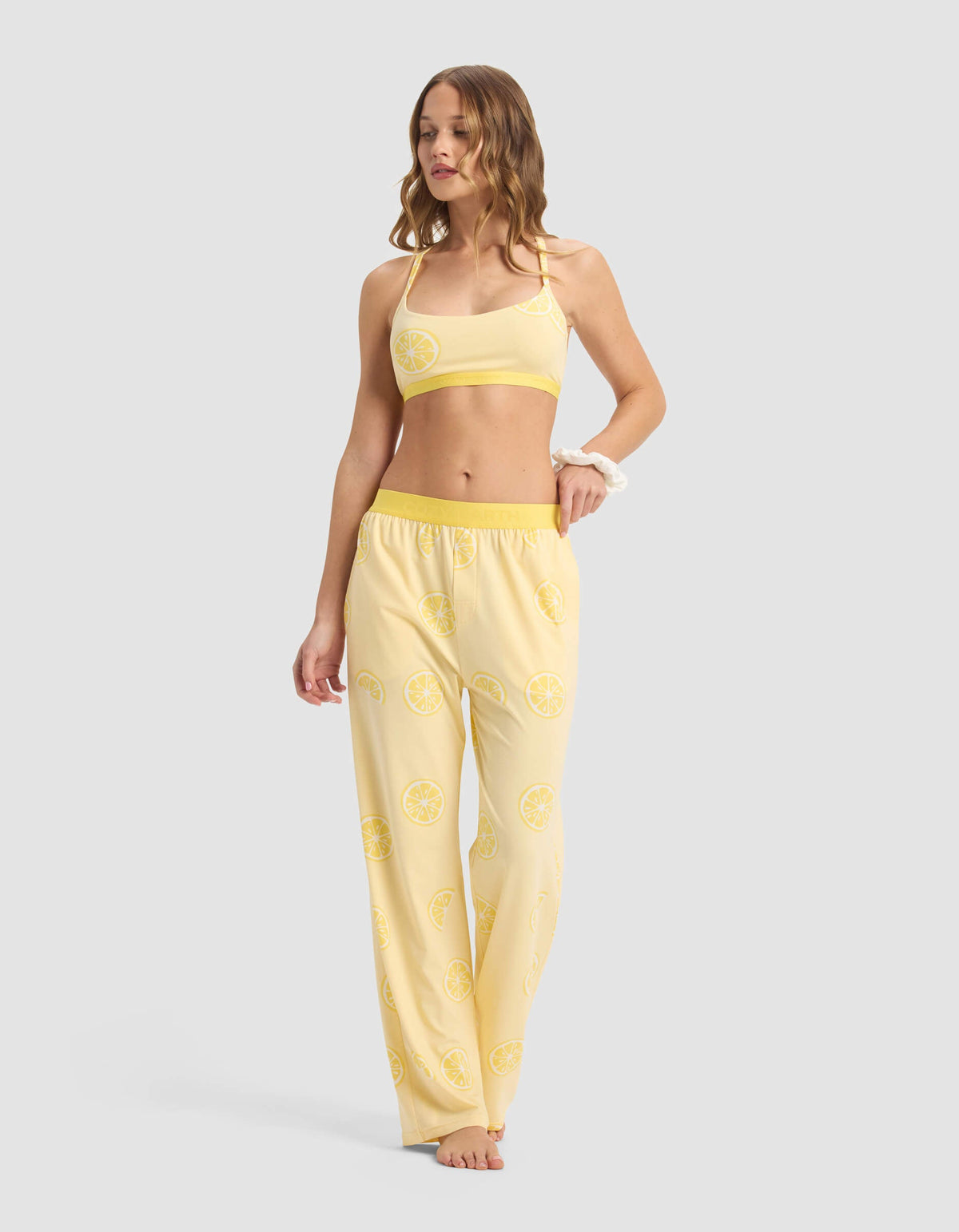 A woman stands barefoot against a plain background wearing Cozy Earth's Women's First Layer Pajama Pant, featuring a yellow color and lemon slice pattern. The set includes a crop top and loose-fitting pajama pants.|Color:Lemons