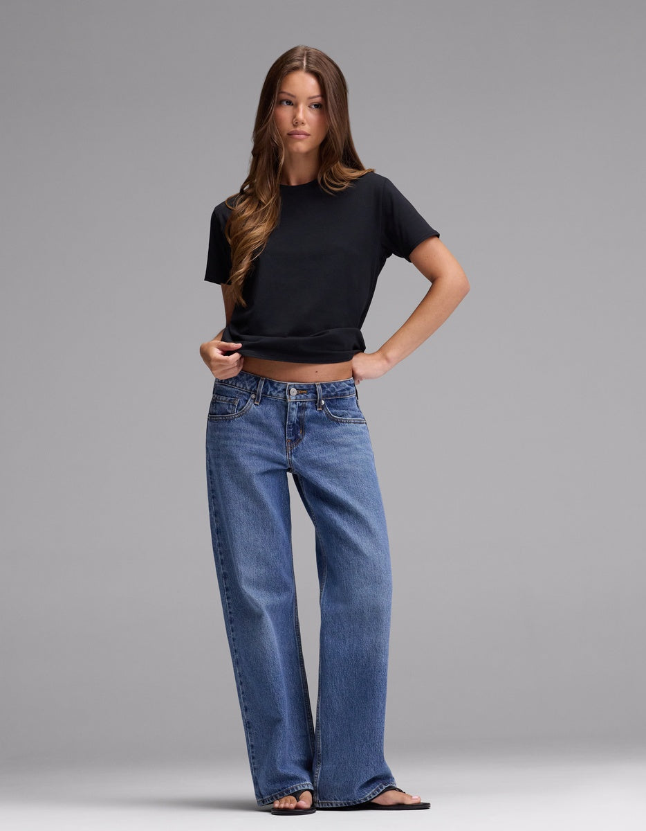 A woman stands against a plain gray background wearing Cozy Earth’s Women’s Go-To Tee in black, paired with blue wide-leg jeans and black sandals. She has long brown hair and a neutral expression, with one hand on her hip.|Color:Jet Black