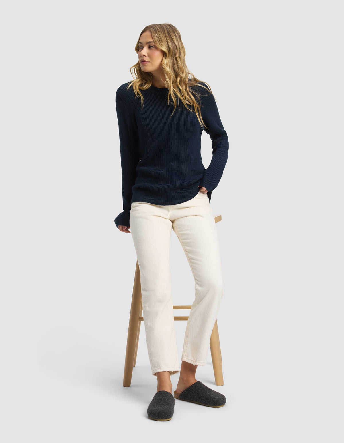 A woman with wavy blonde hair sits on a wooden stool, wearing a navy blue knit sweater, off-white pants, and Cozy Earth's Lakehouse Clog slippers in dark gray, set against a plain light gray background.|Color:Graphite