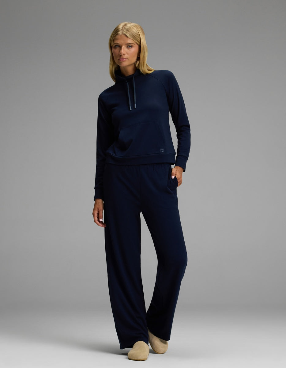 A woman with blonde hair and glasses stands in a relaxed pose against a gray background, dressed in a navy blue hoodie and matching wide-leg pants. She completes her look with Cozy Earth’s Lakehouse Clog in Suede.|Color:Sandalwood