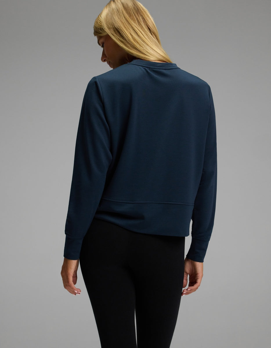 A woman with long blonde hair, seen from behind against a plain gray background, models the Cozy Earth Women’s StudioLite Crewneck in dark blue paired with black leggings.|Color:Eclipse