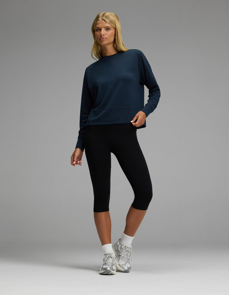 A person stands against a plain gray background, wearing the Cozy Earth Women’s StudioLite Crewneck in navy, paired with black capri leggings, white socks, and gray sneakers. They look slightly to the side with one hand on their hip.|Color:Eclipse