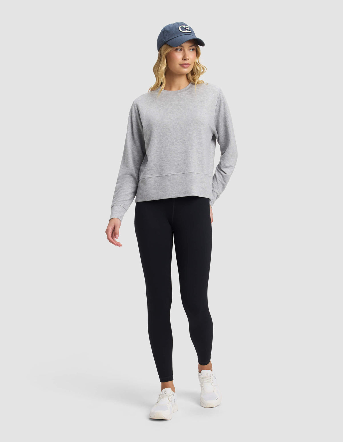 A woman wears the Cozy Earth Women's StudioLite Crewneck, paired with black leggings, white sneakers, and a blue baseball cap. She stands against a light gray background, looking slightly to the side with one hand in her pocket.|Color:Heather Grey