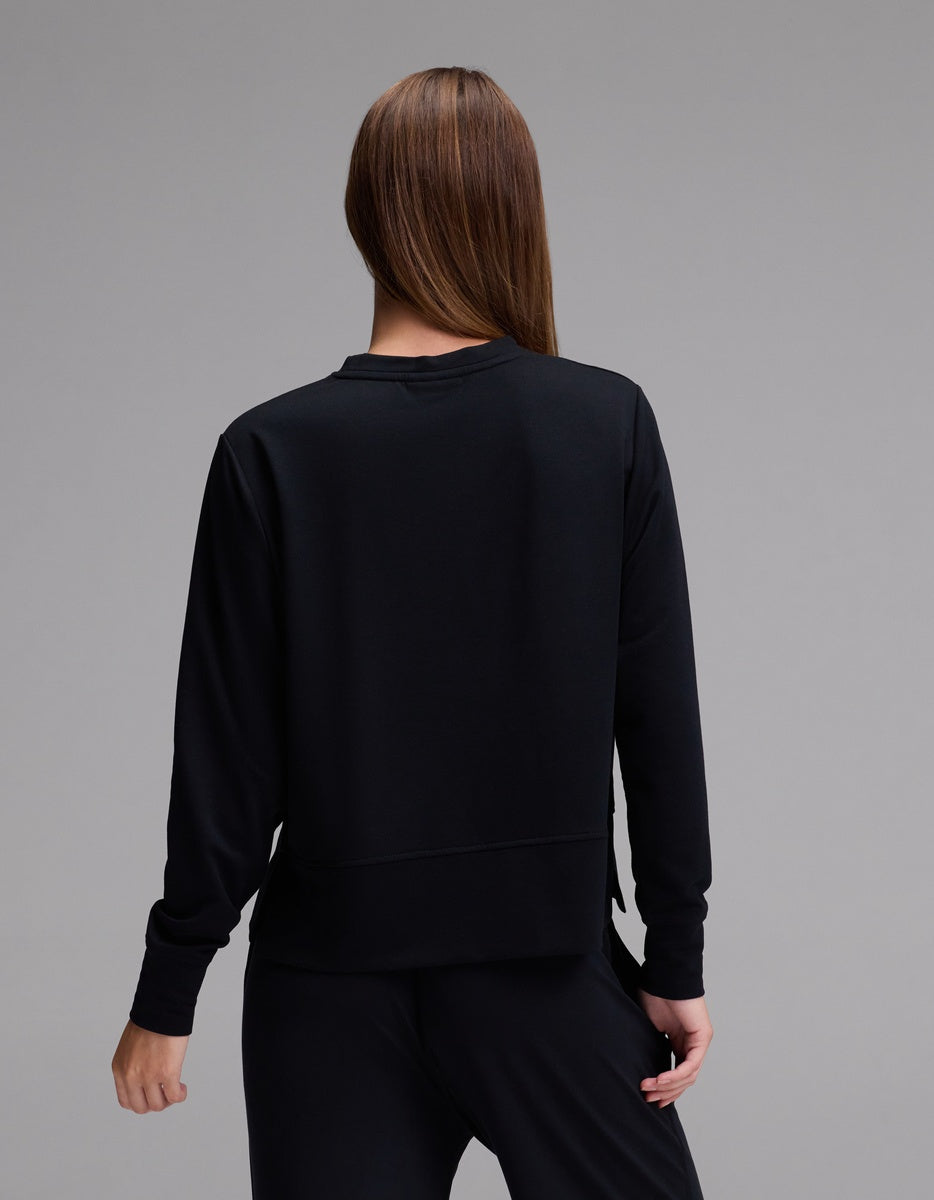 A person with long brown hair, wearing the Cozy Earth Women’s StudioLite Crewneck in black and matching black pants, stands facing away from the camera against a plain gray background.|Color:Jet Black
