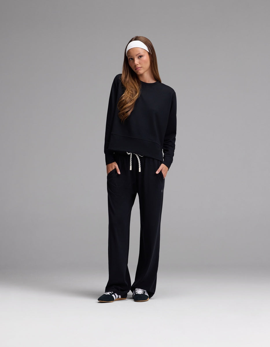 A woman stands against a gray background, wearing the Cozy Earth Women’s StudioLite Crewneck paired with matching black drawstring pants, black sneakers, and a white headband. She has her hands in her pockets and looks straight ahead.|Color:Jet Black
