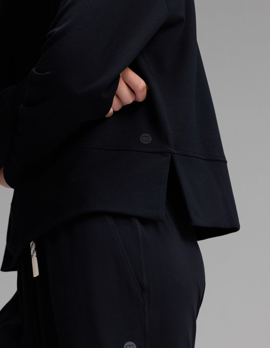 A person wearing the Cozy Earth Women’s StudioLite Crewneck, a loose-fitting black long-sleeve top with a back slit, and matching black pants stands with arms folded against a plain gray background.|Color:Jet Black