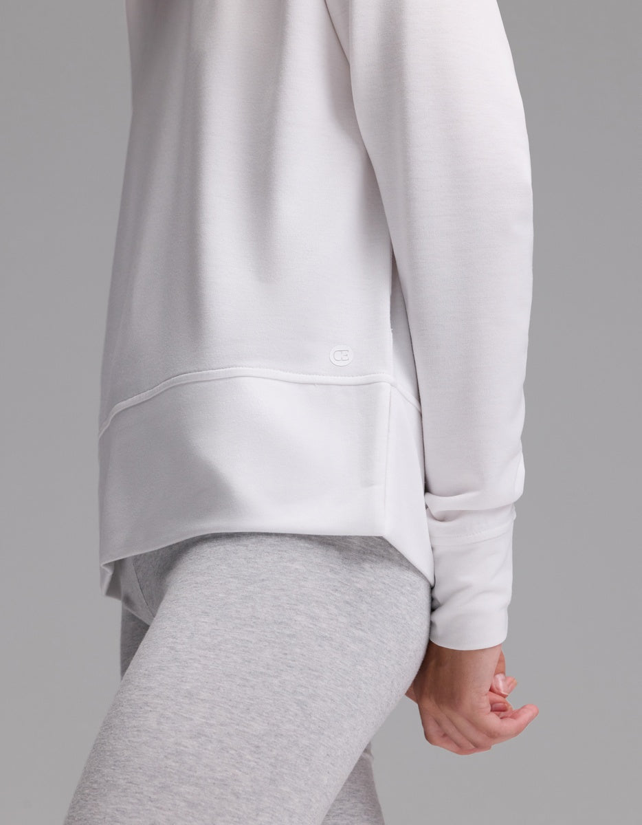 A woman stands sideways against a plain gray background, wearing the Cozy Earth Women’s StudioLite Crewneck in white and light gray leggings, with one arm relaxed at her side.|Color:White