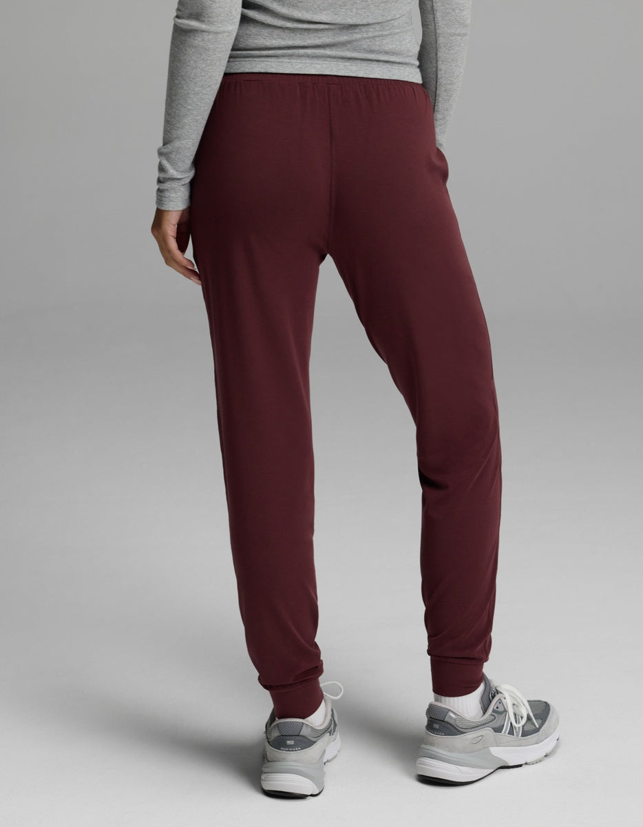 A person is shown from the back, standing on a light gray background, wearing a gray long-sleeve shirt, Cozy Earth Women’s Studio Jogger pants in burgundy, and gray sneakers.|Color:Burgundy