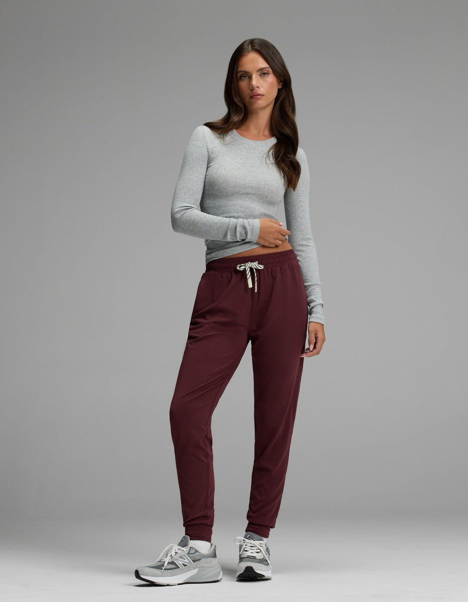 A woman with long brown hair and a neutral expression stands against a gray background wearing the Cozy Earth Women’s Studio Jogger paired with a light gray long-sleeve shirt and gray sneakers.|Color:Burgundy