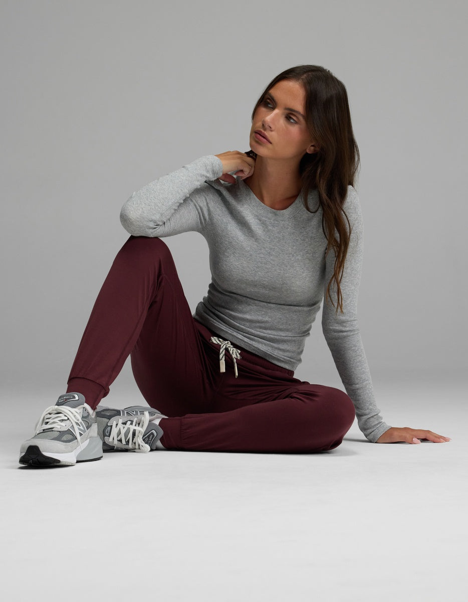 A woman wearing Cozy Earth’s Women’s Studio Jogger in maroon and a gray long-sleeve shirt sits on the floor with one knee up, resting her arm on it and gazing to the side. She finishes her look with gray sneakers against a plain light background.|Color:Burgundy
