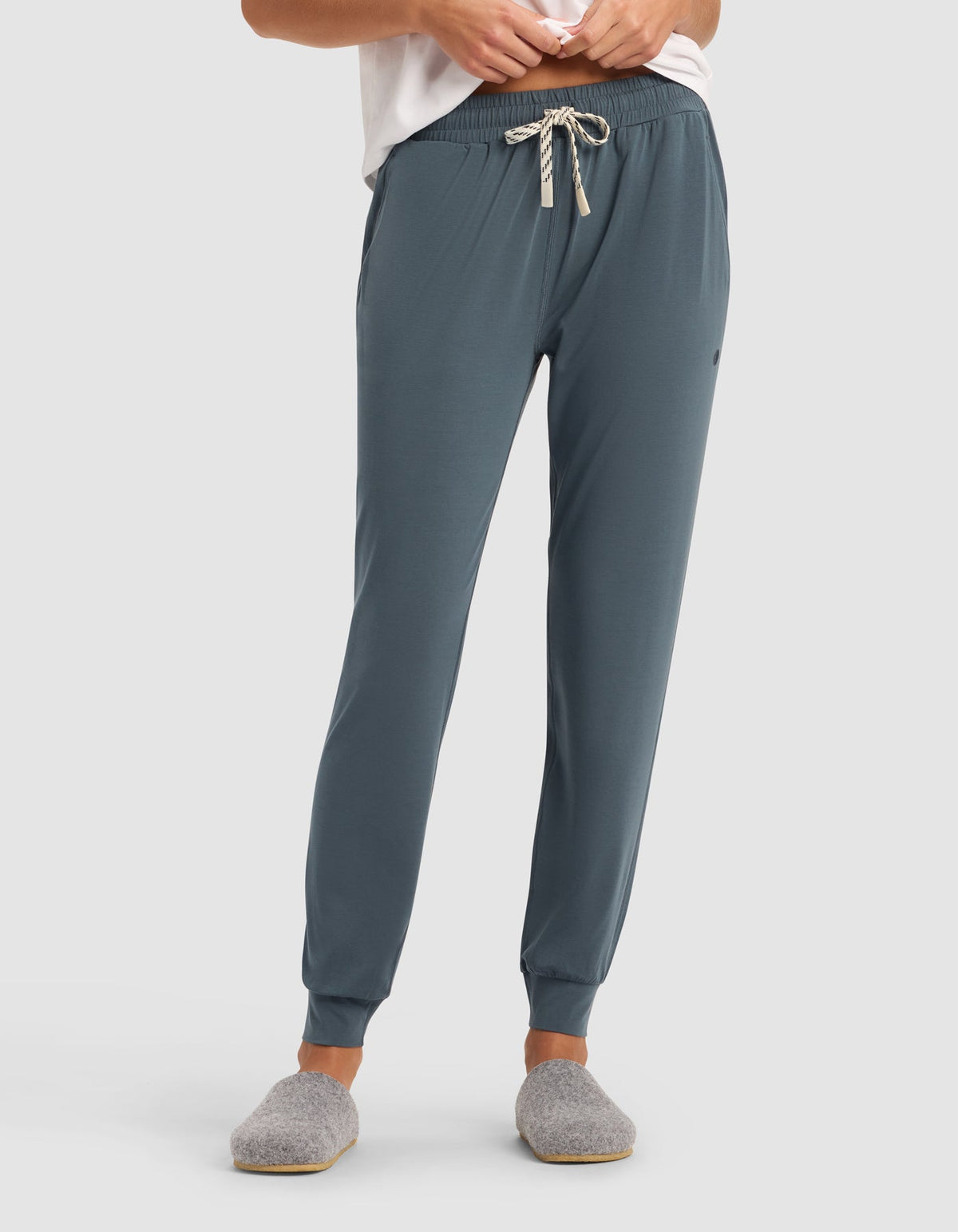 A person wearing Cozy Earth's Women's Studio Jogger in teal with a striped drawstring, paired with a white top and gray slippers, stands against a plain light background. Their face is not visible.|Color:Dawn