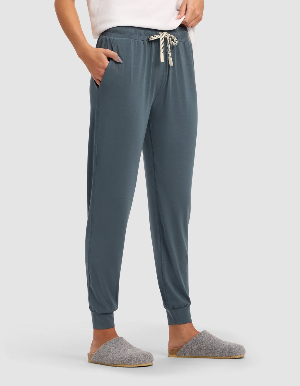 A person stands with hands in pockets, wearing Cozy Earth Women's Studio Jogger in teal with a white drawstring, a white top, and gray slip-on slippers against a plain light background.|Color:Dawn