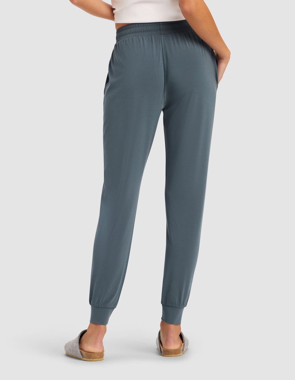 A person in Cozy Earth's Women's Studio Jogger, a white top, and gray slippers stands with their back to the camera, one hand on their hip.|Color:Dawn