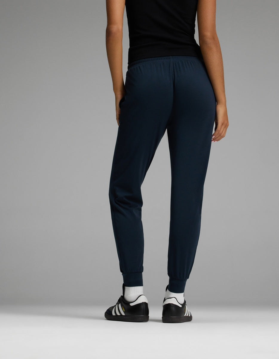 A person wearing a black top, Cozy Earth Women’s Studio Jogger pants in dark blue, and black-and-white athletic shoes stands facing away from the camera against a plain gray background.|Color:Eclipse