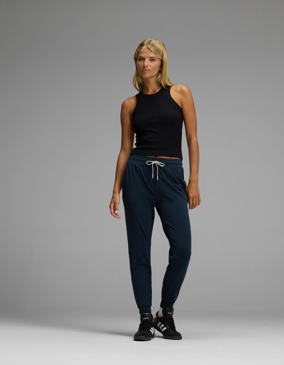 A woman stands against a plain gray background, wearing Cozy Earth’s Women’s Studio Jogger in navy with a drawstring waist, paired with a black sleeveless top and black sneakers.|Color:Eclipse