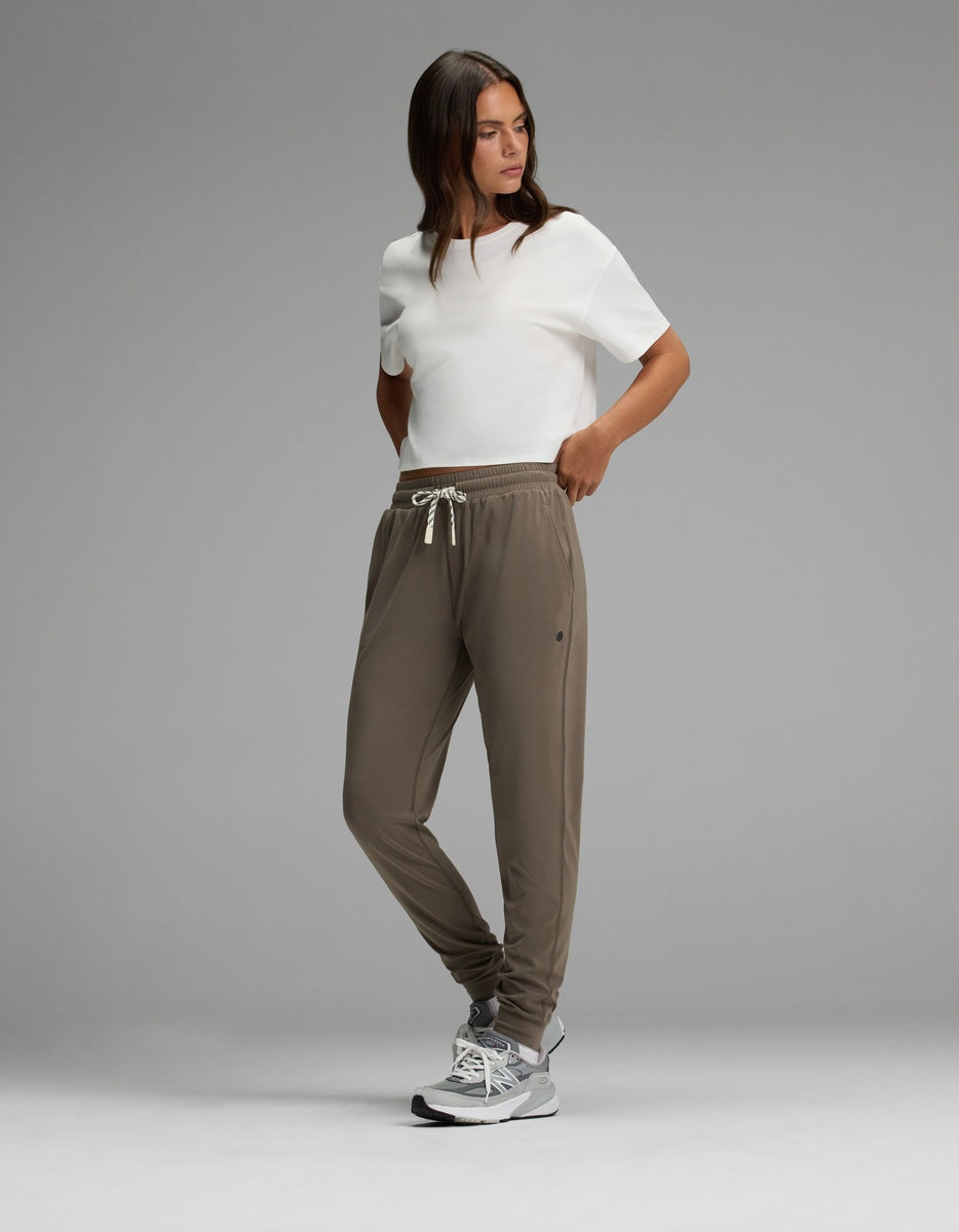 A woman stands against a gray background wearing Cozy Earth’s Women’s Studio Jogger in taupe with a drawstring waist, paired with a white short-sleeve shirt and gray athletic shoes. Her hands are in her pockets and she is looking to the side.|Color:Granite