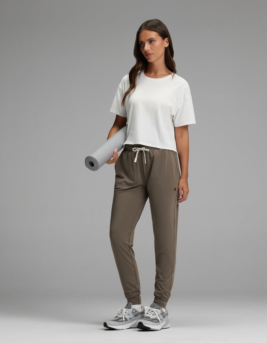 A woman with long brown hair stands on a gray background, wearing Cozy Earth’s Women’s Studio Jogger in taupe and a white T-shirt. She holds a rolled-up yoga mat and wears sneakers.|Color:Granite