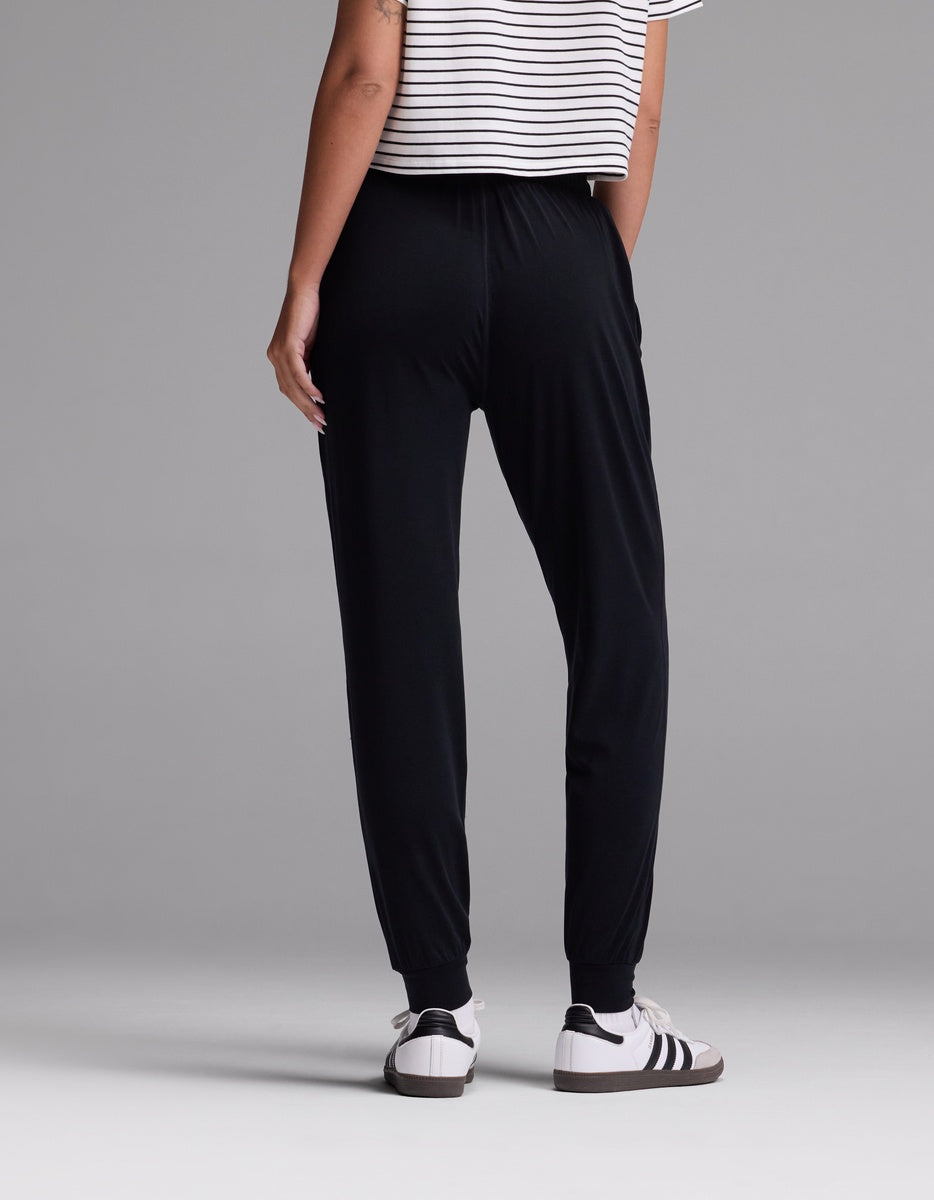 A woman is standing against a gray background, wearing Cozy Earth’s Women’s Studio Jogger pants in black, paired with a white and black striped short-sleeve shirt and white sneakers with black stripes. The photo is taken from the back.|Color:Jet Black
