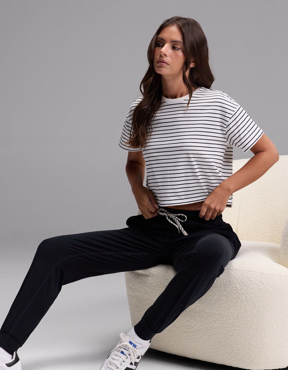 A woman with long brown hair and a relaxed expression sits on a white chair against a plain gray background, wearing Cozy Earth’s Women’s Studio Jogger in black, paired with a black and white striped t-shirt and white sneakers.|Color:Jet Black