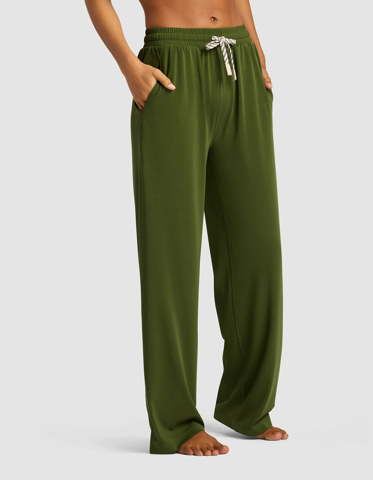A woman models the Cozy Earth Women's Studio Wide Leg Pant in olive green, featuring a white and gray striped drawstring, hands in pockets, barefoot against a light gray background.|Color:Grove