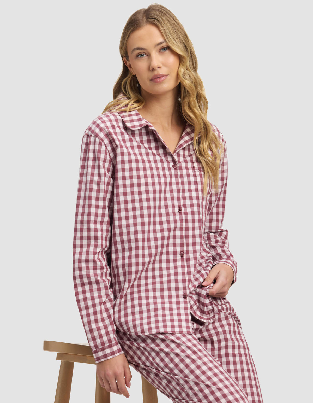 A woman with long blonde hair sits on a wooden stool, wearing the Cozy Earth Women's Sutton Long Sleeve Pajama Top in red and white checks, paired with matching pants, and looks to the side with a neutral expression.|Color:Mulberry Gingham