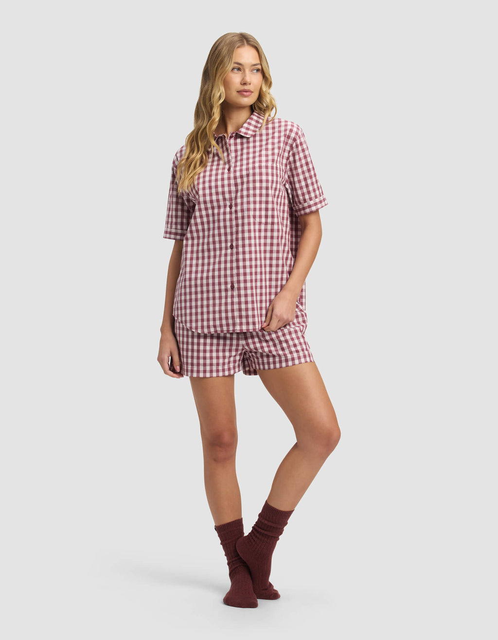 A person wears the Cozy Earth Women's Sutton Short Sleeve Pajama Top in red and white gingham with matching shorts and dark red socks, standing against a plain light gray background.|Color:Mulberry Gingham