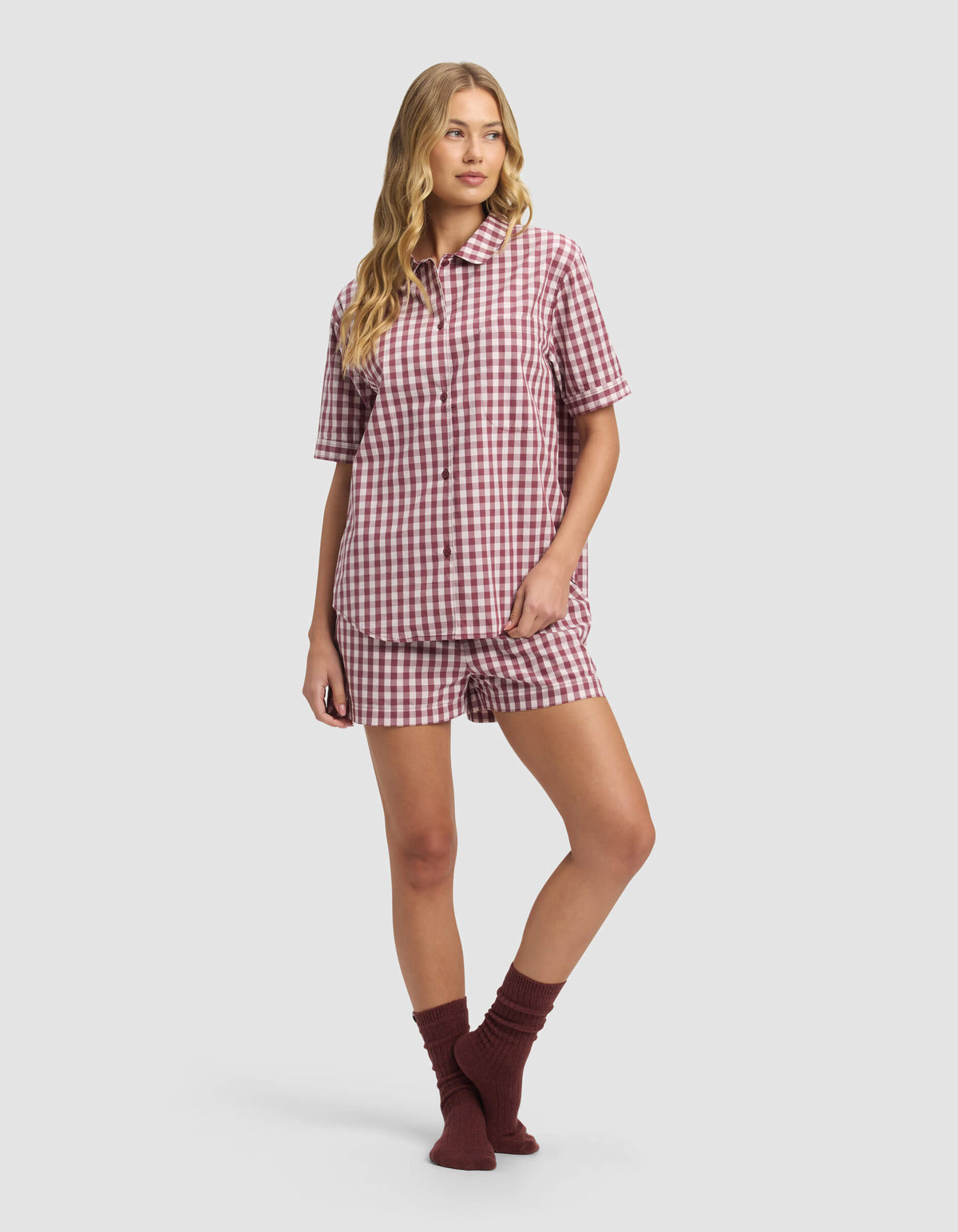 A person wears the Cozy Earth Women's Sutton Short Sleeve Pajama Top in red and white gingham with matching shorts and dark red socks, standing against a plain light gray background.|Color:Mulberry Gingham