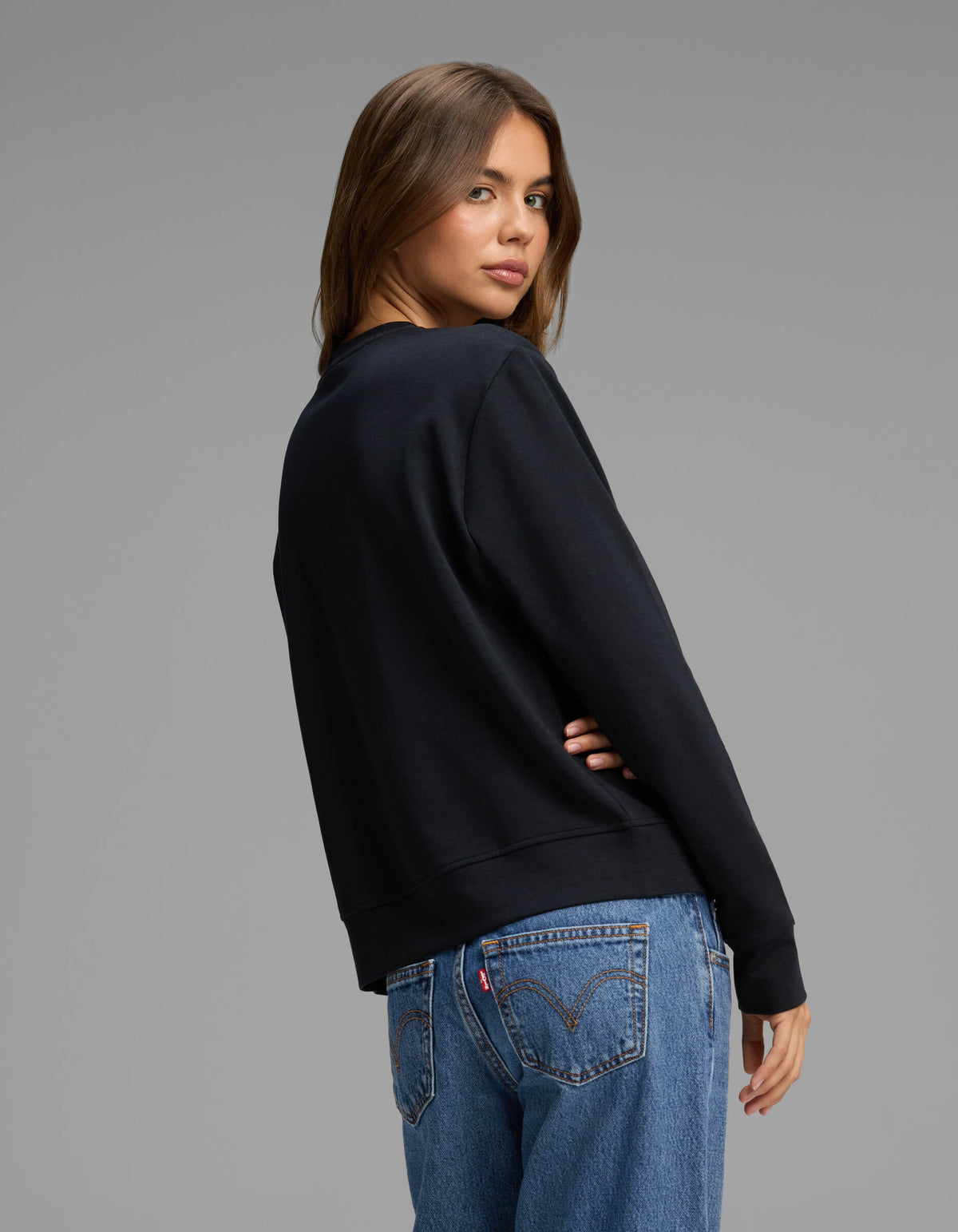 A woman with long brown hair wears the Cozy Earth Women's Versa Crewneck in black and blue jeans, standing with her back turned and head looking over her shoulder against a plain gray background.|Color:Jet Black