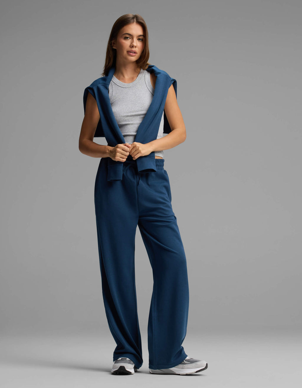 A woman stands against a plain gray background, looking forward with a relaxed posture, wearing Cozy Earth's Women's Versa Straight Leg Pant in blue, paired with a gray tank top and a matching blue sweatshirt draped over her shoulders.|Color:Obsidian