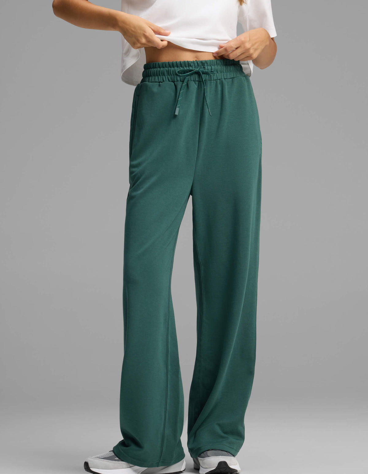 A model showcases the lower half of the Women's Versa Straight Leg Pant by Cozy Earth in green, paired with a white shirt and white sneakers, standing against a plain grey background and seen adjusting the waistband.|Color:Uniform