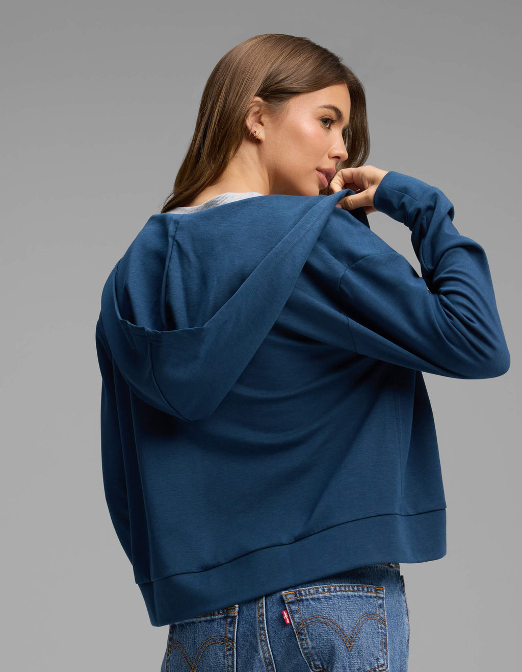 A woman with long brown hair wears the Cozy Earth Women's Versa Full-Zip Hoodie in blue and jeans, looking over her shoulder while holding the collar. She stands against a plain gray background.|Color:Obsidian