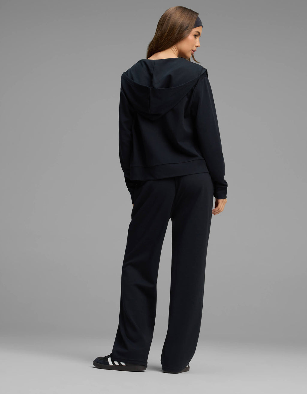 A woman stands facing away, wearing the Cozy Earth Women's Versa Zip-Down Hoodie & Straight Leg Pant Set in black, paired with black slide sandals featuring white stripes, against a plain grey background.|Color:Jet Black