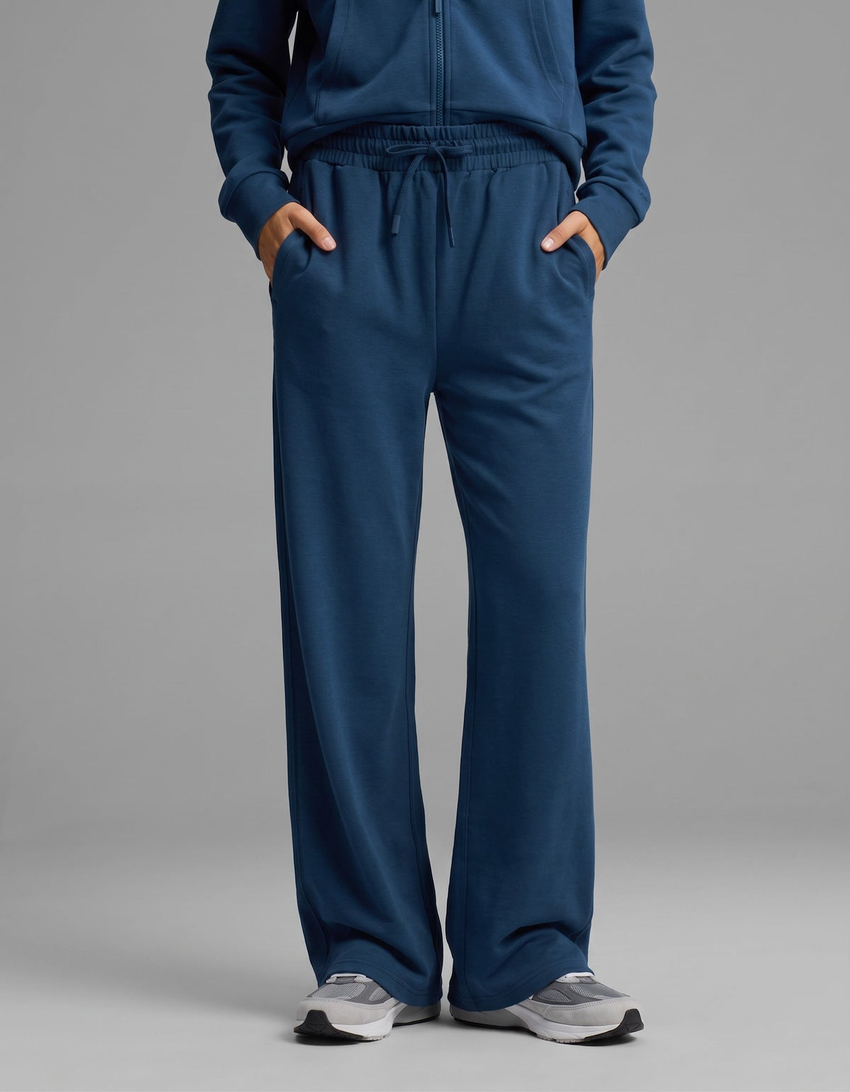 A person models Cozy Earth's Women's Versa Zip-Down Hoodie & Straight Leg Pant Set in blue, paired with gray sneakers, standing against a plain gray background with hands in pockets.|Color:Obsidian