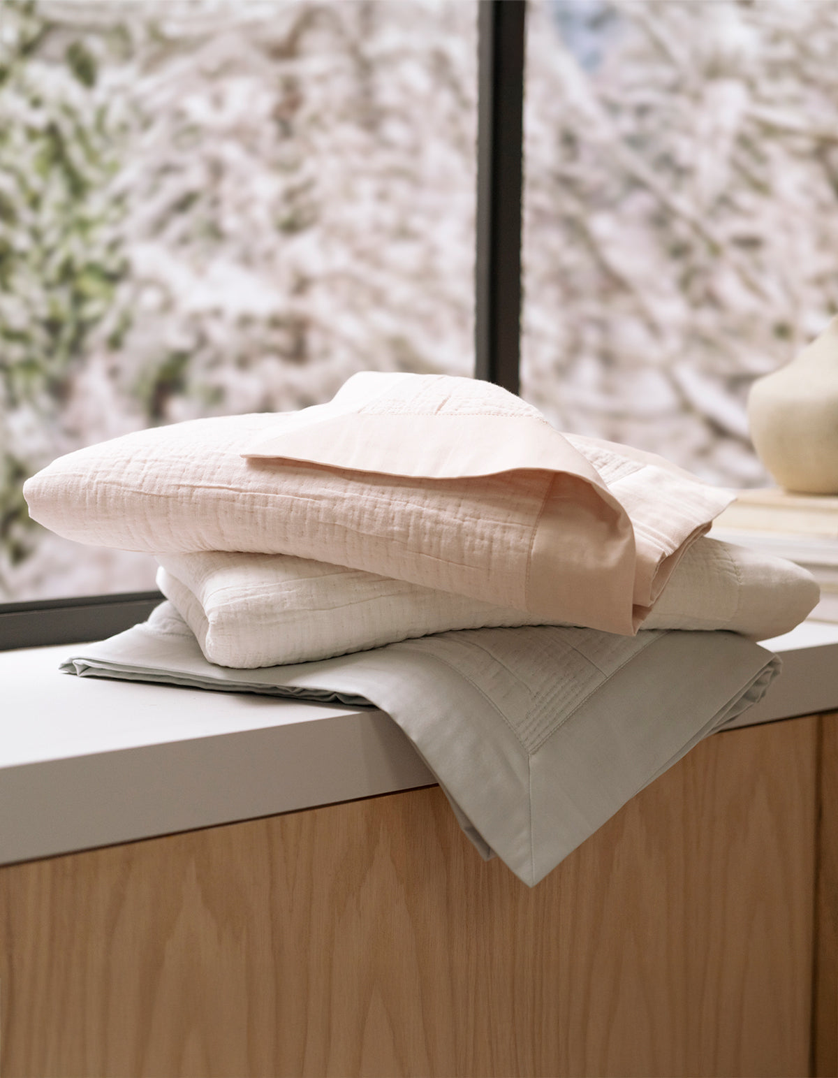 Three Cozy Earth Little Bee Matelasse Baby Blankets in soft pastels are neatly folded and stacked on a light wooden surface by a window, with blurred snow-covered trees visible outside.|Color:Sea Salt