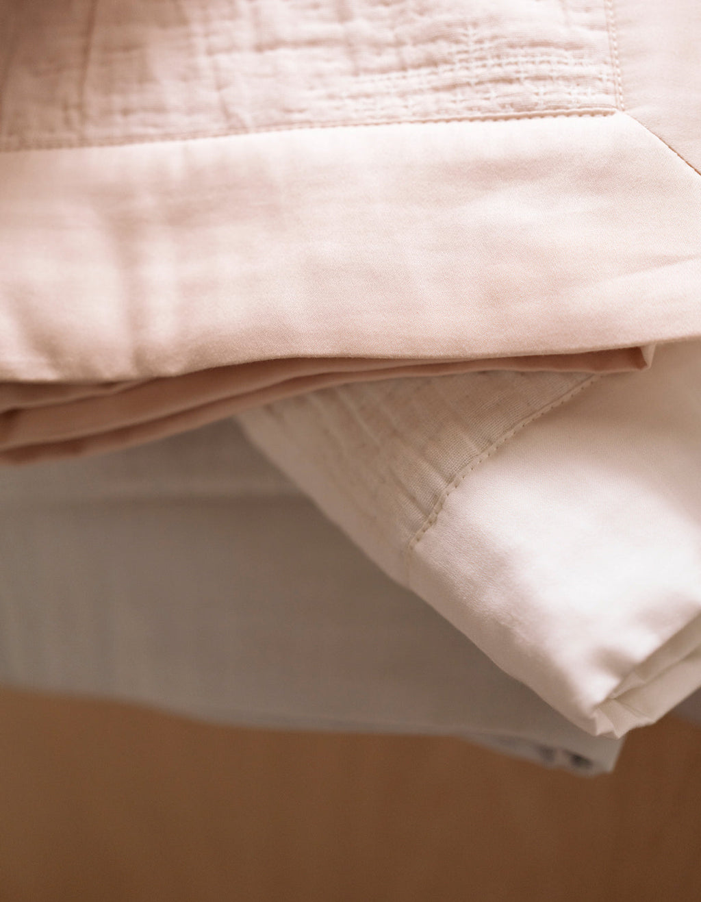 A close-up of the Cozy Earth Little Bee Matelasse Baby Blanket shows soft pink and white fabrics neatly folded in a stack, featuring smooth, slightly textured materials.|Color:Vapor