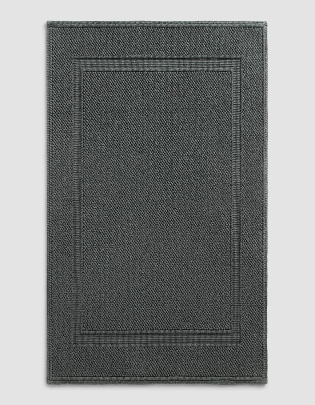 The Cozy Earth Classic Bath Mat features a dark gray, rectangular design with a textured surface and double border, shown against a plain light background.|Color:Graphite