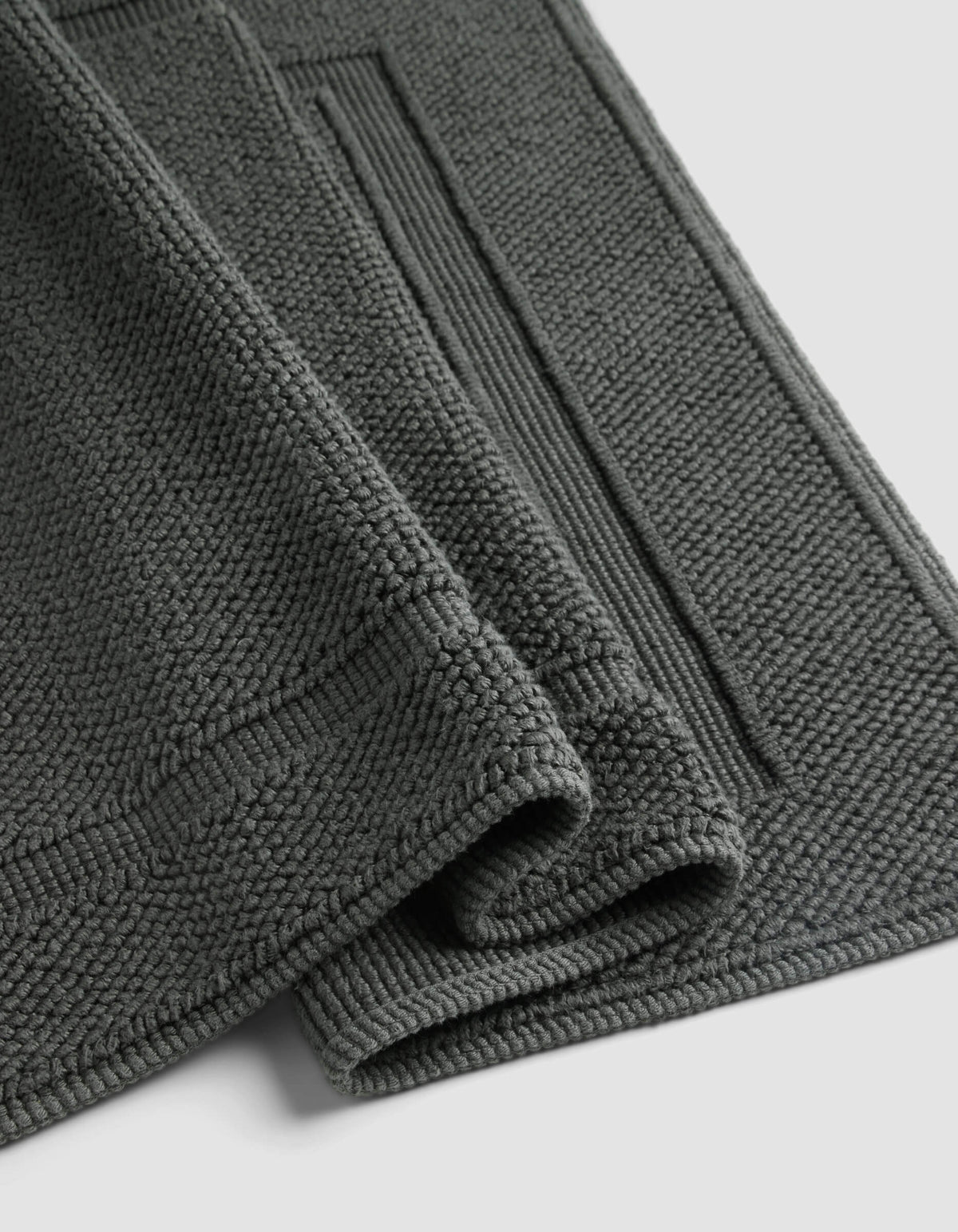 The Cozy Earth Classic Bath Mat, shown folded against a light background, features a dark grey textured finish with a thick, soft pile and a rectangular border pattern.|Color:Graphite