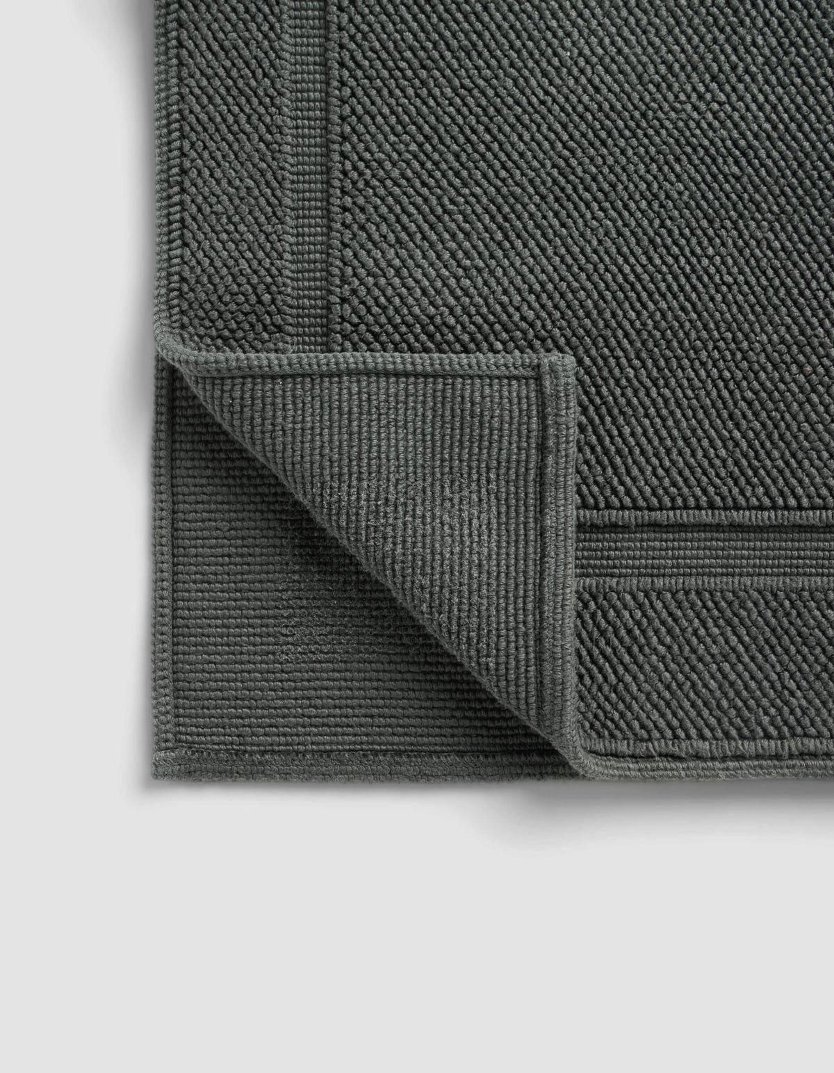 The Cozy Earth Classic Bath Mat, in dark gray with a textured woven pattern and bordered edges, is displayed on a light background with one corner folded to reveal the underside.|Color:Graphite