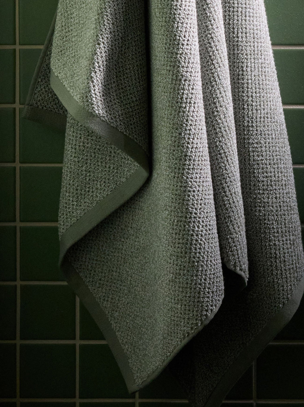 A Nantucket Bath Sheet in Heathered Sage is pictured hanging against a green tiled wall. |Color:Heathered Sage